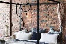 a simple industrial bedroom with red brick walls, a black metal frame bed, wooden and wicker furniture and cozy textiles