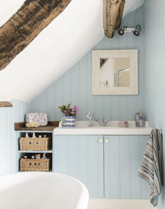 a small powder blue attic bathroom with wooden beams, baskets for storage and a free-standing tub