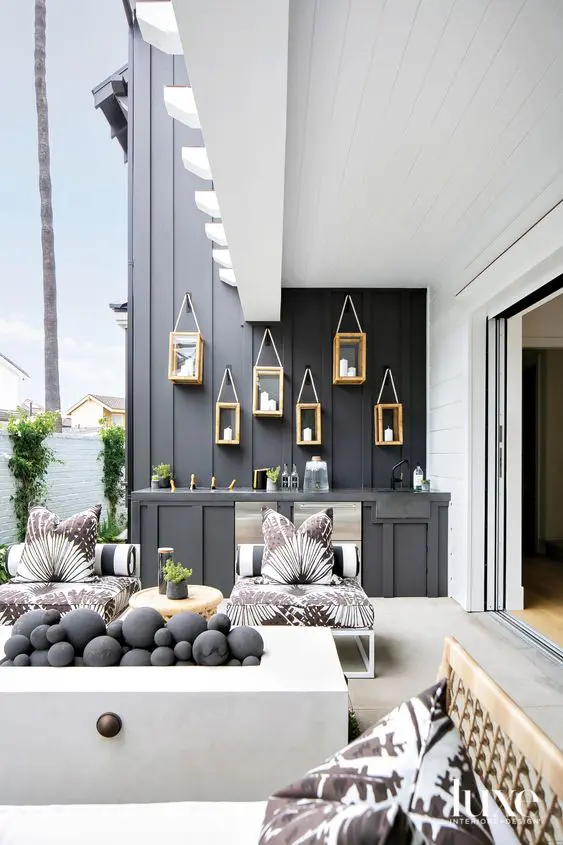 a stylish contemporary outdoor space with a black wall and a bar counter, white chairs with printed upholstery, a white fireplace wiht black pebbles