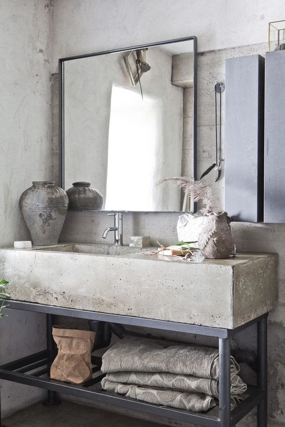 A wabi sabi meets industrial bathroom with concrete walls, a concrete vanity with metal legs, mirrors and concrete vases