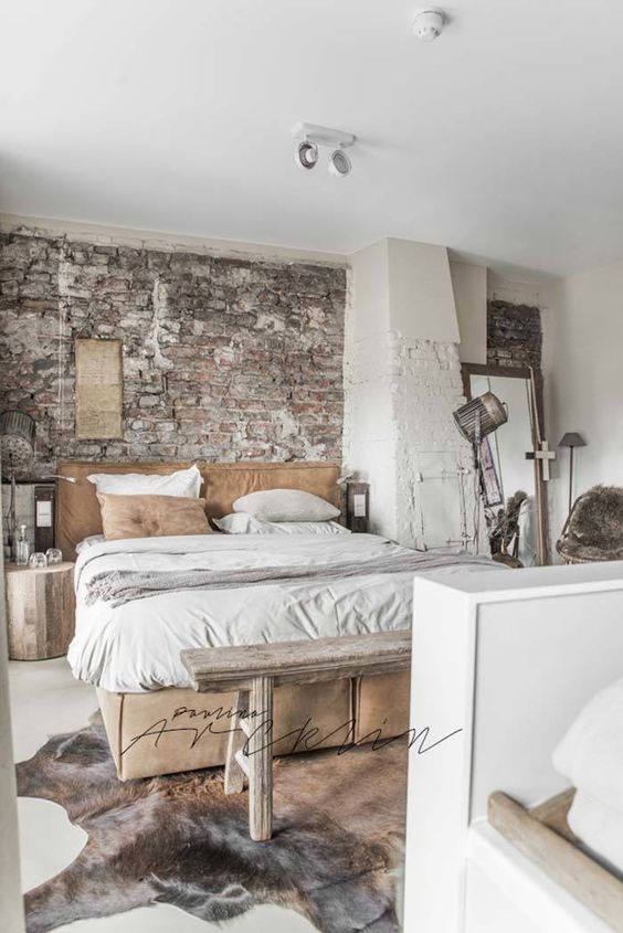 a white industrial meets wabi-sabi bedroom with a brick wall, a leather bed with neutral bedding, a vintage hearth, lamps and tree stumps as nightstands