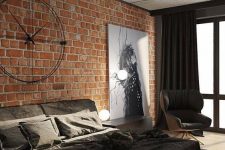 an elegant industrial bedroom with a red brick accent wall, a black upholstered bed with black bedding, a creative metal clock and lights