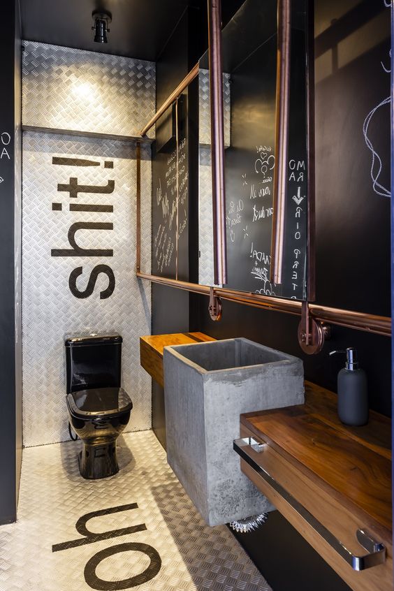 an industrial bathroom with black walls, white tiles, a wooden vanity, a concrete sink, a mirror in a copper frame and a black toilet