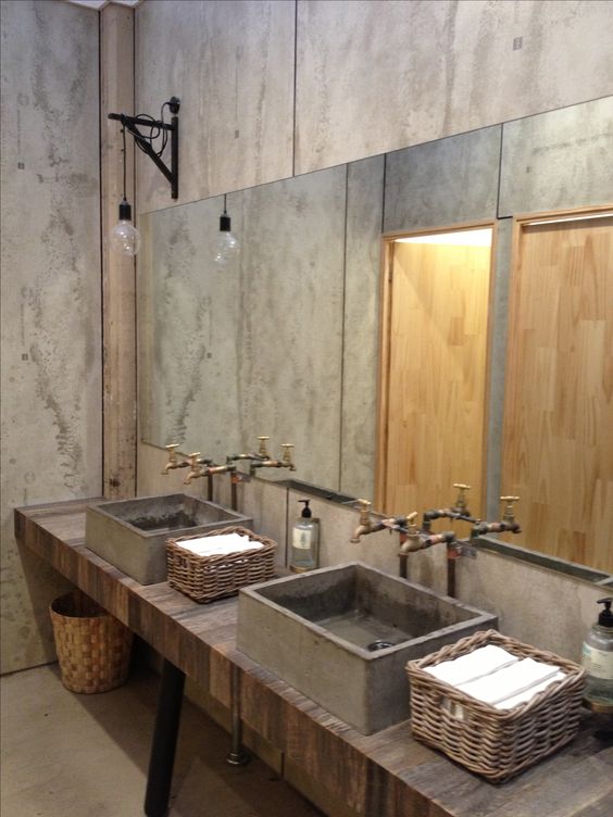 an industrial bathroom with concrete walls, a wooden vanity, concrete sinks and a large mirror plus vintage exposed pipes