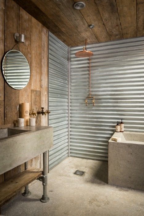 an industrial bathroom with rough wood and corrugated steel, a stone vanity and bathtub, exposed pipes and lamps