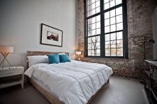 an industrial bedroom with a shabby chic brick wall, a wooden bed with neutral bedding, a white nightstand, double-height windows for more light