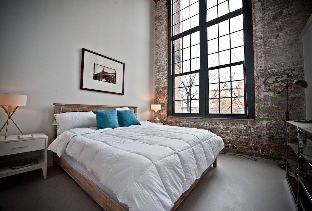 An industrial bedroom with a shabby chic brick wall, a wooden bed with neutral bedding, a white nightstand, double height windows for more light