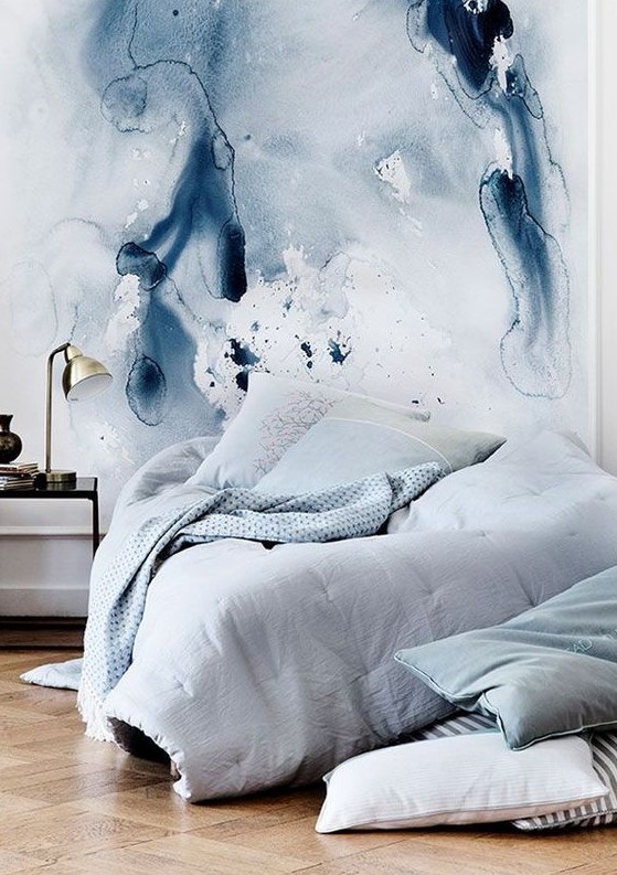 an ocean-inspired bedroom with a blue watercolor wall, a bed with grey and light blue bedding, a black side table and a gold table lamp