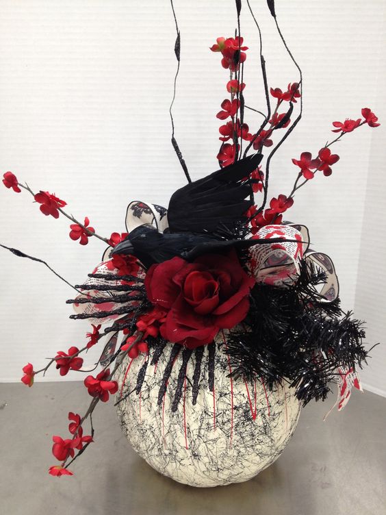 a Halloween centerpiece of a painted pumpkin, black glitter skeleton hands, red blooming branches, blooms and a crow on top