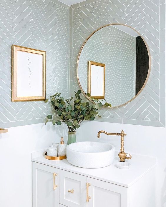 a beautiful bathroom with pale blue chevron tiles, a white vanity, gold handles, a round mirror in a gold frame, gold fixtures and an artwork in a gold frame
