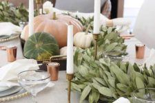 a chic modern Thanksgiving tablescape with a lush greenery runner, natural pumpkins and tall candles plus touches of copper
