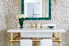 a glam bathroom with Dolmatin walls, a gold sink stand, gold sconces and a mirror in a beautiful green frame plus a green vase