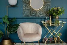 a glam space with a teal wall and paneling, a creamy chair, a gold bar cart, a gold planter, a mirror in a beautiful gold burst frame and a floor lamp