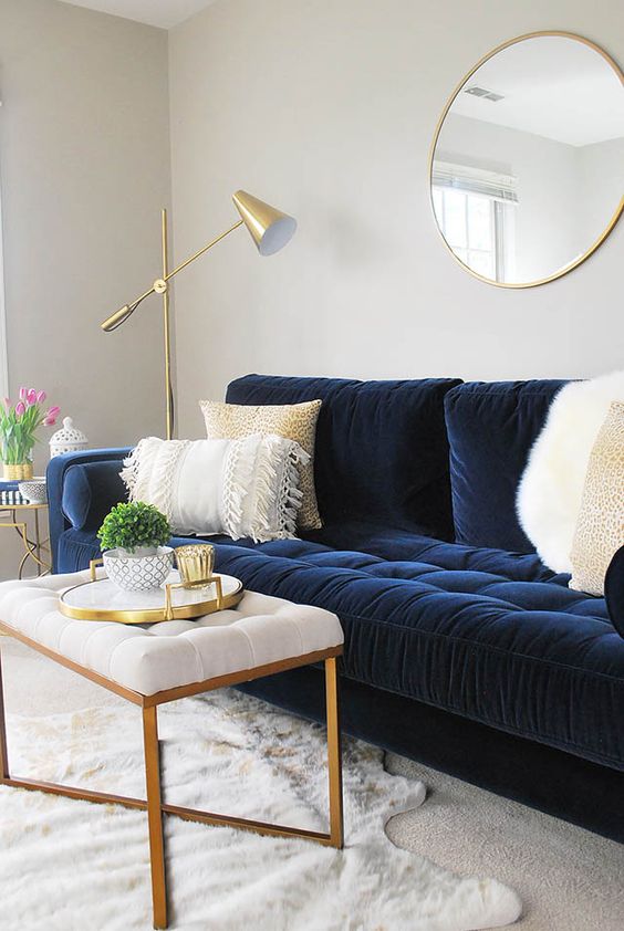 a modern living room in neutrals with a navy velvet sofa, a white pouf with gold legs, a gold floor lamp and a round mirror