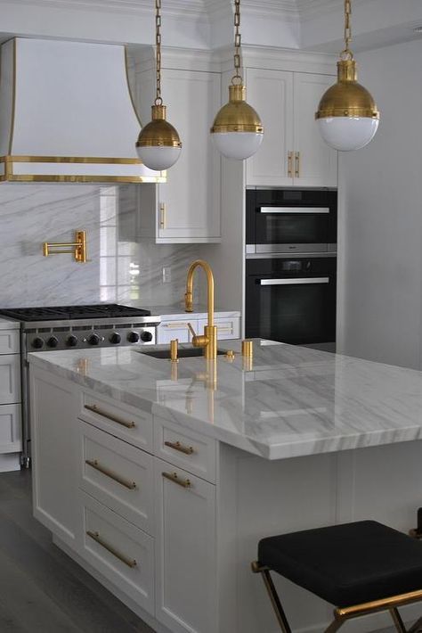 a modern white kitchen with white stone countertops and a backsplash, gold fixtures and a hood lined with gold, gold pendant lamps