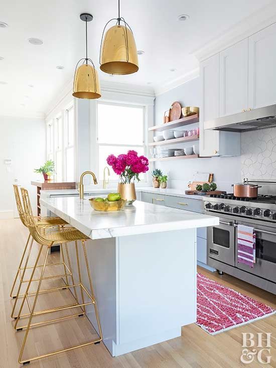 a pretty coastal kitchen done with pale blue cabinets,, gold fixtures, gold pendant lamps and tall stools, a pink rug and pink blooms
