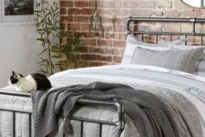 a pretty industrial bedroom with a red brick accent wall, a metal piping bed with grey bedding, pendant lamps and potted plants