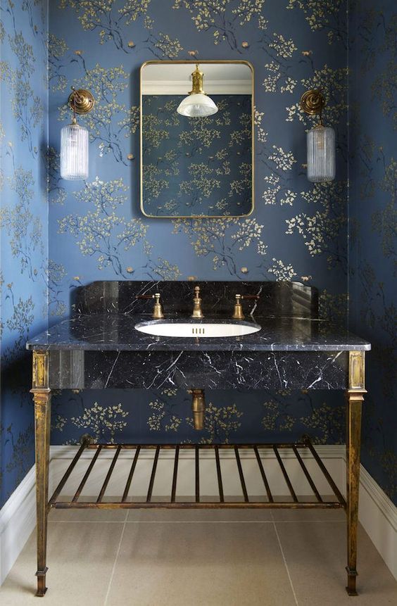 a refined bathroom with navy and gold floral wallpaper, a black marble sink on gold legs and gold fixtures, a mirror in a gold mirror and gold sconces