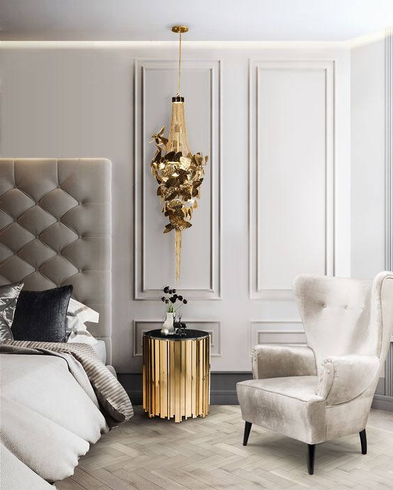 a refined bedroom done in creamy and dove grey, with a bed with an oversized headboard, a wingback chair, a gold nightstand and a gold butterfly pendant