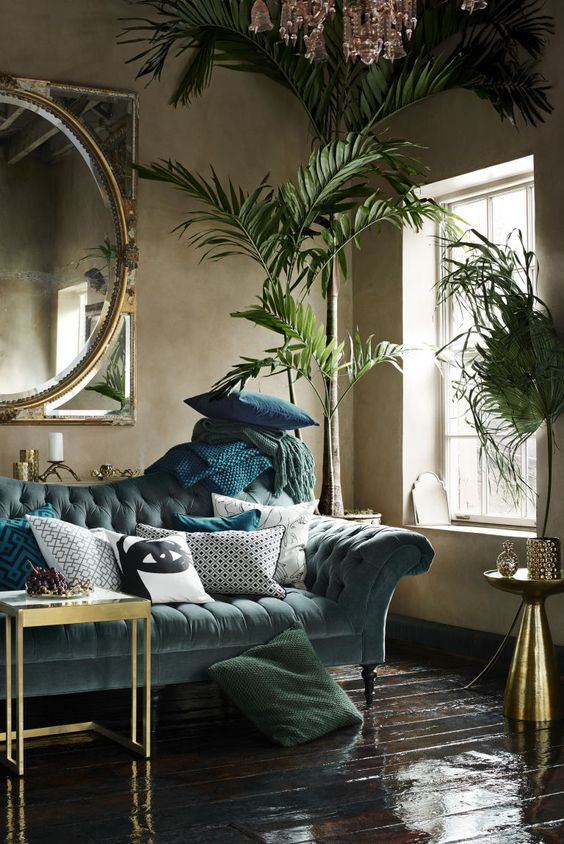 a refined living room with a moody feel, a pale blue sofa, green and blue textiles, potted plants, gilded coffee tables, a crystal chandelier and a large mirror in a gilded frame