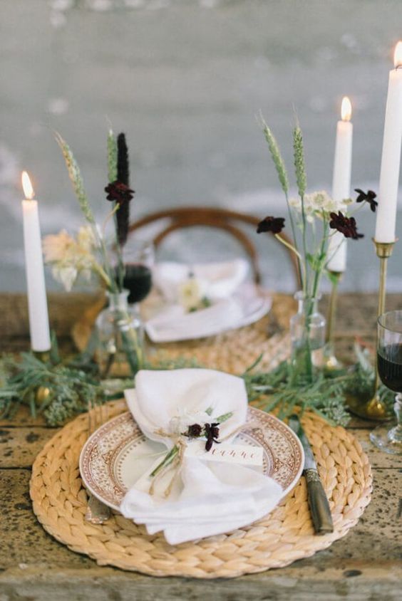 a simple Thanksgiving table with woven placemats, tall candles, dark blooms and lots of greenery