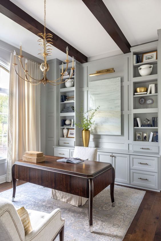 A sophisticated vintage inspired home office with light grey cabinets, a dark stained desk, delicate gold touches   a chandelier, a lamp, a vase and pillows