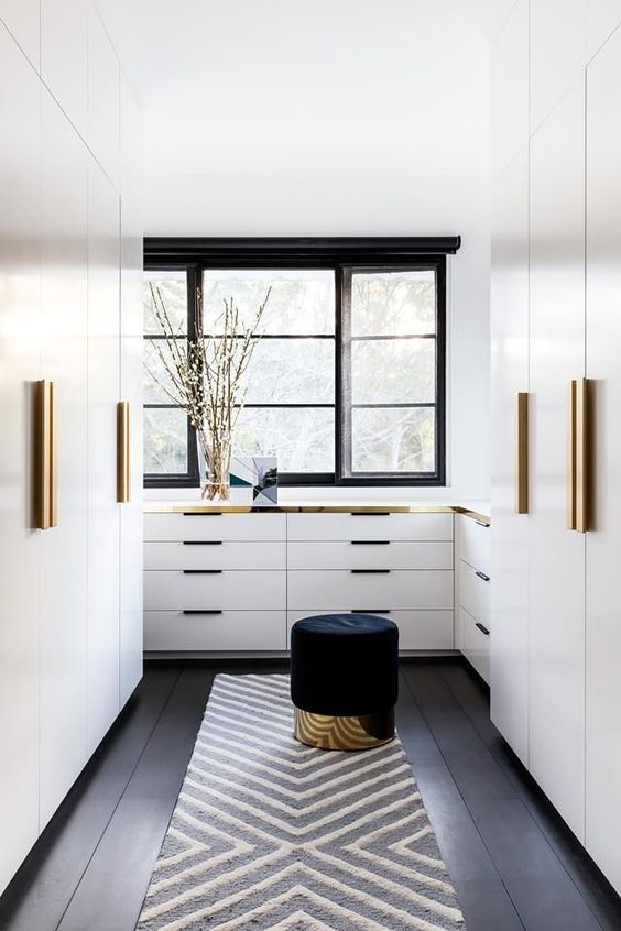 a stylish modern closet with white wardrobes and dressers with gold handles and gold touches here and there, a black pouf with a gold base
