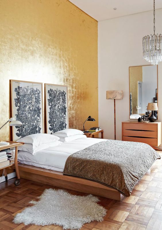 an eclectic bedroom with a shiny gold accent wall, a simple bed with catchy art, a matching dresser and cart nightstands