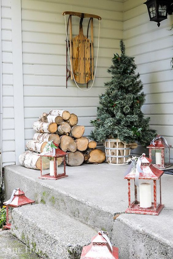 a Christmas tree in a basket with burlap, firewood, red candle lanterns and a sleigh will make your front porch very cozy and inviting