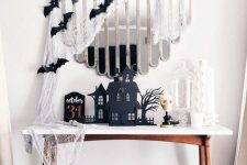 a Halloween console table with cheesecloth, black bats, black houses and a white skull is a bold and cool idea