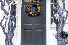 a Halloween porch with black spiderwebs, skeletons and skulls, a black and orange pumpkin wreath on the door