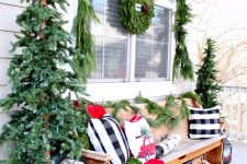 a bright rustic Christmas porch with two trees in buckets, a candle lantern, a fir wreath and a garland and bright festive pillows