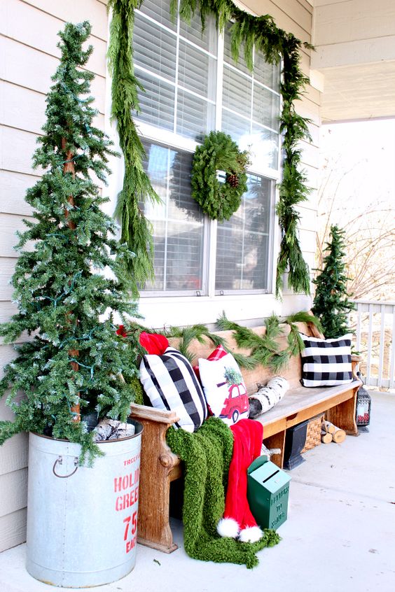 a bright rustic Christmas porch with two trees in buckets, a candle lantern, a fir wreath and a garland and bright festive pillows