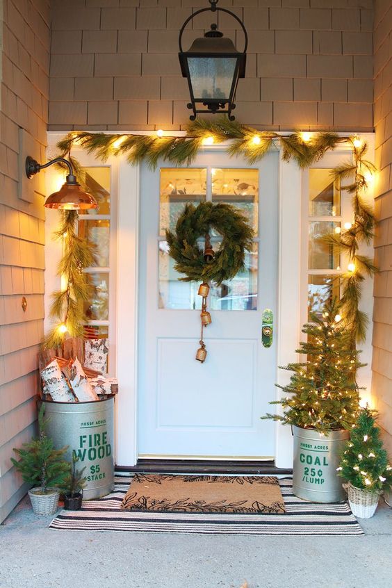 a bucket with firewood, a bucket with a Christmas tree and a mini one next to it, a fir garland, a fir wreath with bells
