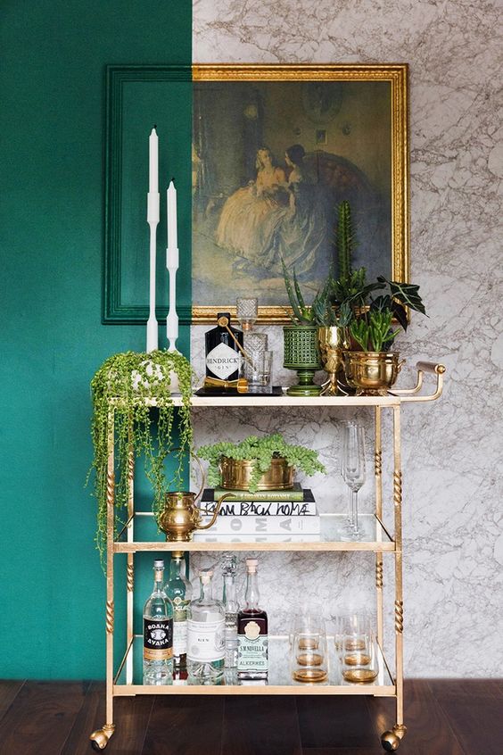 a chic and glam gold bar cart on casters, with potted greenery and succulents, with books and glasses plus a vintage artwork over the cart