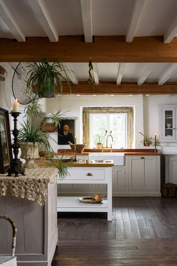 a cottage kitchen with wooden beams, white shaker cabinets and butcherblock countertops, some art and lots of potted plants