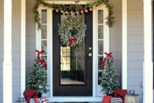 a cozy Christmas front porch with an evergreen and pinecone garland, a snowy wreath, mini trees with lights and lots of gift boxes