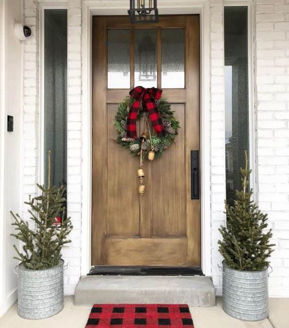 a duo of Christmas trees in buckets, a Christmas wreath with bells and a plaid bow make the space very cozy