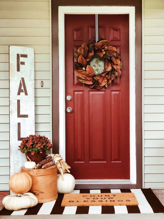 a fall porch with a bold leaf wreath on the door, fresh pumpkins, corn and corn husks and soem blooms in a pot for Thanksgiving