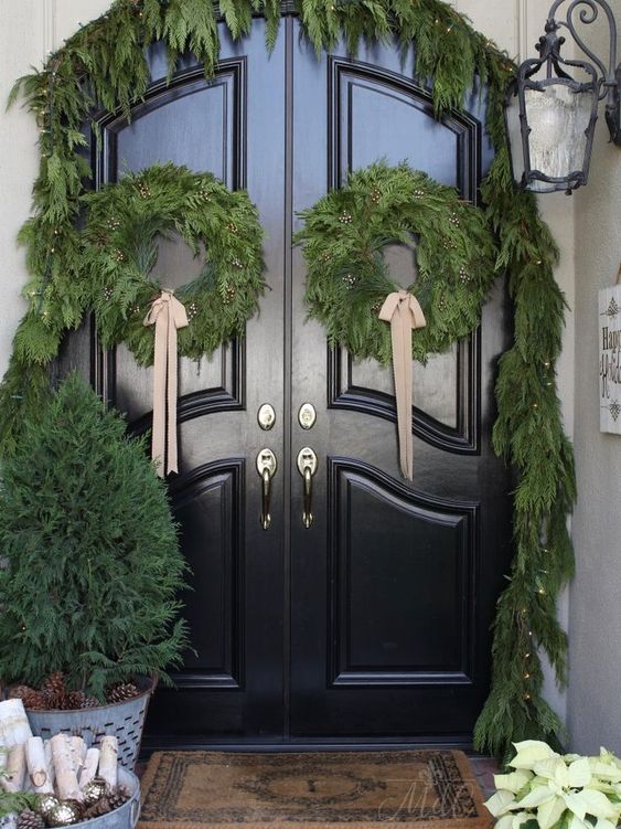 a farmhouse Christmas front door with an evergreen garland with lights, matching wreaths with bows and a mini tree in a bucket