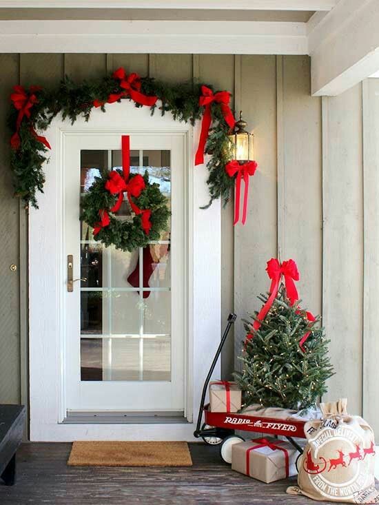 a farmhouse Christmas porch with an evergreen garland with red bows and a matching wreath plus a tree with lights and gift boxes
