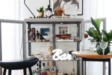 a glass bar cabinet with artworks, a lamp, some plants and a neon sign is a very chic idea