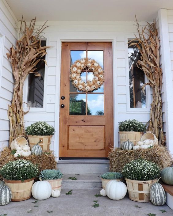 a gorgeous Thanksgiving porch with a white pumpkin wreath and corn husks, fall blooms and natural pumpkins in baskets