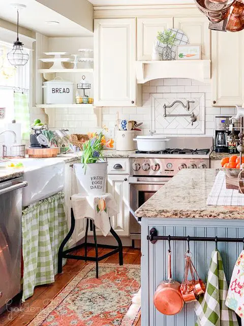 a little and cute cottage kitchen with white cabinets, a blue kitchen island and stone countertops, pendant lamps and printed textiles