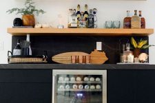 a modern home bar with sleek black cabinets, a wine cooler, open shelves, potted greenery, wine bottles and wine glasses