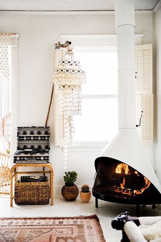 a neutral boho living room with a white Malm fireplace, woven chandeliers and curtains, a printed box, potted plants and a printed rug