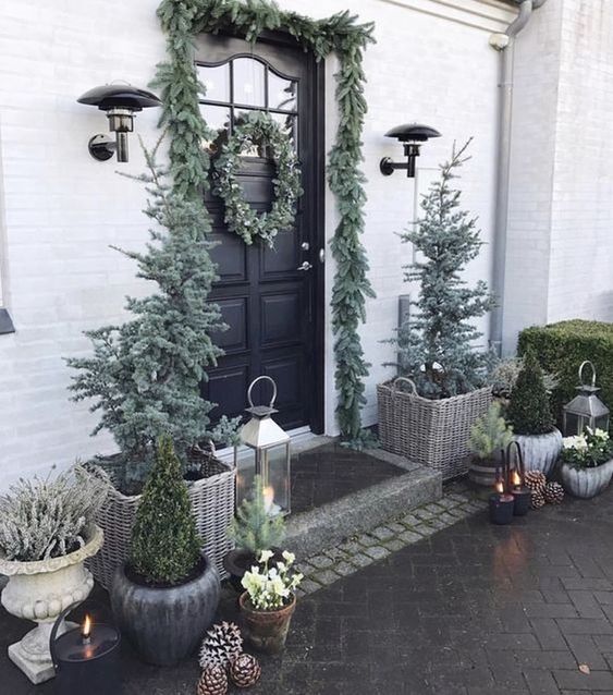 a pale winter front door with an evergreen garland and wreaths, Christmas trees, candle lanterns and pinecones