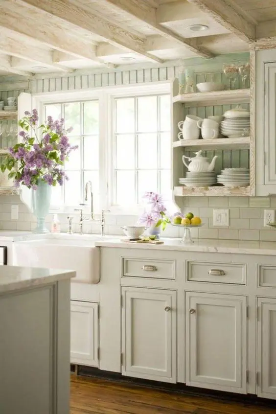 a pastel cottage kitchen with light green shaker cabinets, a beadboard backsplash, open shelves and neutral stone countertops