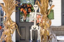 a rustic Halloween porch done with stacked pumpkins, corn husks, a catchy green, orange and black wreath and a skeleton in rubber boots