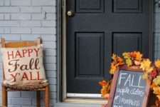 a sign decorated with fall leaves, a crate with blooms and a pumpkin, a fun pillow and a plywood pumpkin sign on the door
